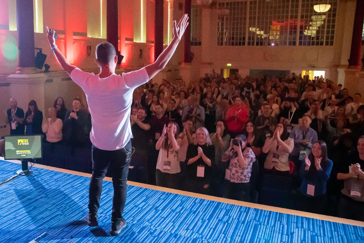 A speaker on stage, arms raised, facing a packed hall on its feet